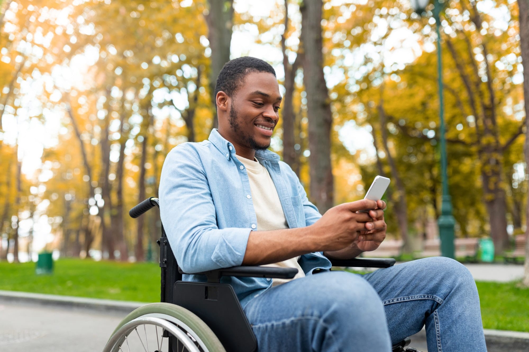Cheery disabled black guy in wheelchair using mobile phone, checking email or messages at autumn
