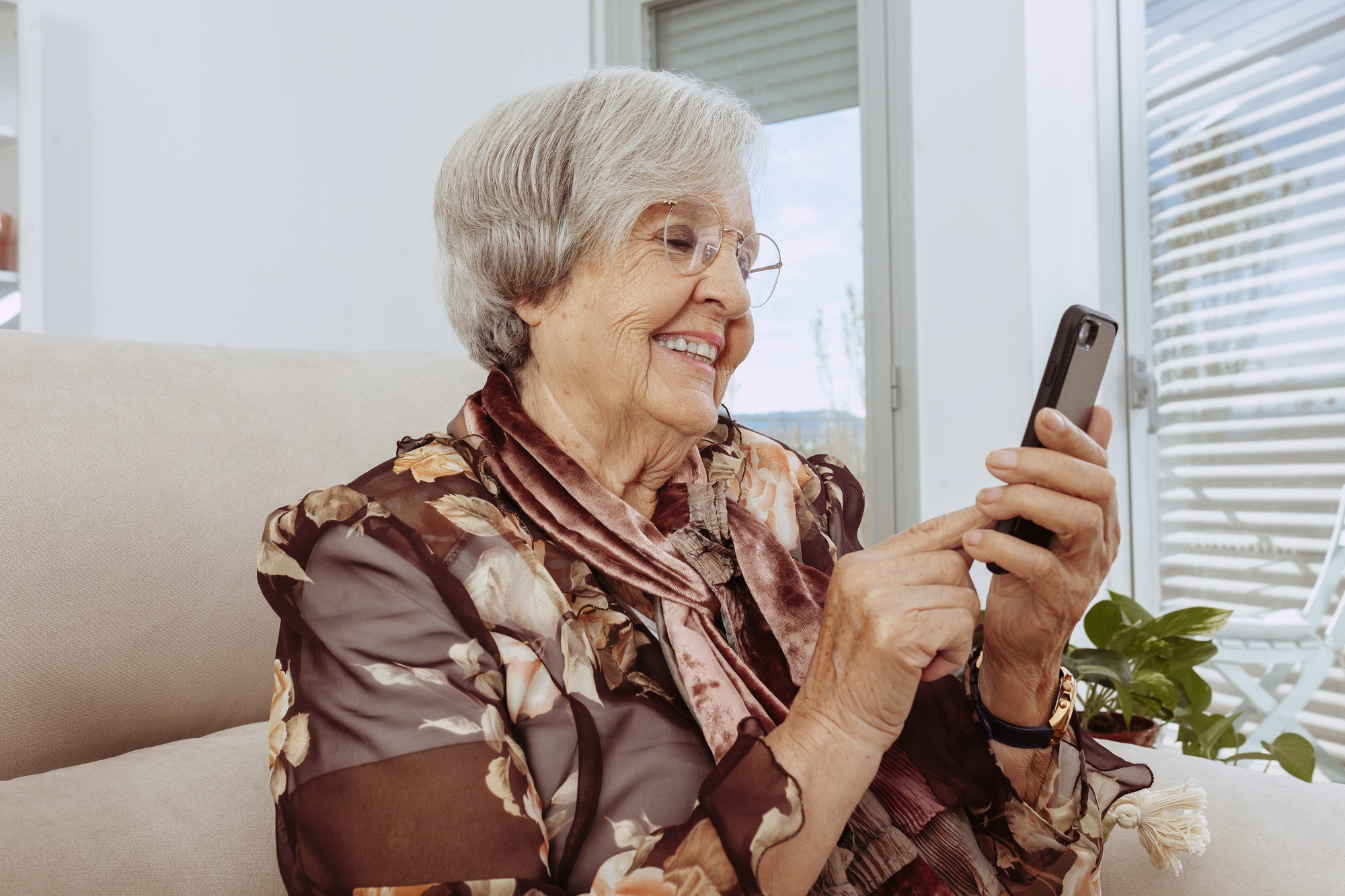 Old grandmother using smartphone sitting on couch at home